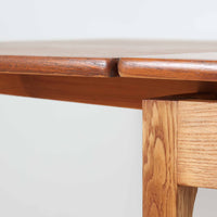 Ejner Larsen & Aksel Bender Madsenが手掛け、Naestved Mobelfabrikで製作されたエクステンションダイニングテーブルは、チーク材の天板とオーク材の脚部を組み合わせた北欧ビンテージらしい上質な佇まいが魅力です。直線的で端正なフォルムの中に、天板縁の丁寧な面取りや幕板の滑らかな造形など、当時の高い職人技が随所に感じられます。幅120cmから最大230cmまで伸長可能な構造により、日常使いから来客時まで柔軟に対応でき、空間や人数に合わせた使い方が可能です。経年変化によって深みを増した木肌の表情が、長く愛用されてきたビンテージならではの存在感を放ち、ダイニングの主役として落ち着きと品格を添えてくれる一台です。脚先には床へのダメージや汚れを防ぐためにチーク材がジョイントされており、実用性を考慮した当時の設計思想がうかがえます。また、裏面には「SKM」の刻印が確認でき、スウェーデン軍(Svenska KrigsMakten)の備品として使用されていたことを示す、背景のある希少な個体です。