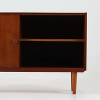 Domino Mobler社製のサイドボードは、北欧デンマークのビンテージ家具らしいシンプルかつ機能的なデザインが魅力です。チーク材を用いた温かみのある木目と深みのある色調が空間に落ち着きを与えます。左側には4杯の引き出し、右側にはスライド扉付きの収納棚を備え、食器や日用品をすっきり収めることが可能です。特徴的なスクエア型の木製ハンドルや細身の脚部が洗練された印象を与え、ダイニングやリビングでの収納家具としてだけでなく、テレビボードや飾り棚としても活躍する実用性と存在感を兼ね備えています。年月を経た風合いがより一層の魅力を引き立てる逸品です。