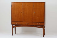 FDBモブラーでBorge mogensenの後任としてアートディレクターを務めたPoul m. Voltherによる、美しく洗練されたキャビネット。良質なマホガニー材の本体に、ビーチ材の脚部を組み合わせたコントラストが魅力です。真鍮の丸い鍵穴が特徴的な3枚の扉を備え、中央には調節可能な棚、両側には精巧な蟻継ぎが施された移動式の引き出しを配しています。下部には真鍮の俵型ハンドル付きの引き出しが3杯並び、細部へのこだわりが際立ちます。また、室内で靴を履く文化に配慮し、脚先には汚れ防止のための濃い塗装が施されています。1950年代にデザインされた本作は、先細りの脚やストレッチャーがミニマルな美しさを際立たせる一方、シンメトリーなフォルムや真鍮の装飾が伝統的なエレガンスを醸し出し、モダンとクラシックが調和した逸品です。