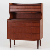 デンマーク製のチーク材によるライティングビューロです。幅85cmと程よいサイズ感で、上段には小引き出しを備え、小物や文具を整理しやすい構造になっています。中央のライティング天板は手前にスライドさせることで作業スペースが確保でき、書き物やノートPCの使用に最適です。さらに、内部には鏡が備わっており、ドレッサーとしても活用できる実用的なデザインです。下段には容量の大きな引き出しが3杯あり、衣類や書類の収納にも対応します。チーク材特有の温かみのある木目と経年による艶が味わい深く、実用性と美観を兼ね備えたビンテージ家具です。