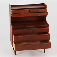 デンマーク製のチーク材によるライティングビューロです。幅85cmと程よいサイズ感で、上段には小引き出しを備え、小物や文具を整理しやすい構造になっています。中央のライティング天板は手前にスライドさせることで作業スペースが確保でき、書き物やノートPCの使用に最適です。さらに、内部には鏡が備わっており、ドレッサーとしても活用できる実用的なデザインです。下段には容量の大きな引き出しが3杯あり、衣類や書類の収納にも対応します。チーク材特有の温かみのある木目と経年による艶が味わい深く、実用性と美観を兼ね備えたビンテージ家具です。