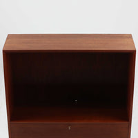 Hans J. Wegnerがデザインし、Ry Mobler社より製造されたこちらのディスプレイシェルフは、チーク材の上質な素材使いと真鍮パーツのディテールが際立つ北欧ビンテージ家具です。シンプルな直線的フォルムの中に、扉付き収納、ドロップダウン式のビューロスペース、小引き出しや仕切り棚など機能的な構造が備わっており、書斎でのデスク兼収納やリビングの飾り棚として多用途に活躍します。年月を経て深みを増した木肌が空間に落ち着いた存在感を与える逸品です。