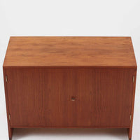 Hans J. Wegnerがデザインし、Ry Mobler社より製造されたこちらのディスプレイシェルフは、チーク材の上質な素材使いと真鍮パーツのディテールが際立つ北欧ビンテージ家具です。シンプルな直線的フォルムの中に、扉付き収納、ドロップダウン式のビューロスペース、小引き出しや仕切り棚など機能的な構造が備わっており、書斎でのデスク兼収納やリビングの飾り棚として多用途に活躍します。年月を経て深みを増した木肌が空間に落ち着いた存在感を与える逸品です。