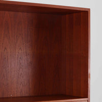 Hans J. Wegnerがデザインし、Ry Mobler社より製造されたこちらのディスプレイシェルフは、チーク材の上質な素材使いと真鍮パーツのディテールが際立つ北欧ビンテージ家具です。シンプルな直線的フォルムの中に、扉付き収納、ドロップダウン式のビューロスペース、小引き出しや仕切り棚など機能的な構造が備わっており、書斎でのデスク兼収納やリビングの飾り棚として多用途に活躍します。年月を経て深みを増した木肌が空間に落ち着いた存在感を与える逸品です。