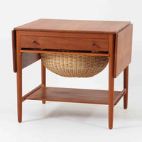 Hans J. Wegnerがデザインし、Andreas Tuck社から発表されたAT33ソーイングテーブルは、チーク無垢材を贅沢に用いた希少なモデルです。両側のバタフライ式天板で作業スペースを拡張でき、仕切り付きの引き出しとラタン製バスケットが裁縫道具や小物を美しく収納します。下段の棚板も備えた実用性の高い構造で、繊細な造作と職人技が光る北欧ビンテージ家具の逸品です。
