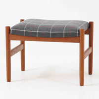 デンマーク製のこちらのスツールは、チーク材の温かみあるフレームに、日本メーカーのチェック柄ファブリックで張り替えを施した、北欧ビンテージ家具らしい洗練と実用性を兼ね備えた一脚です。脚部は丸棒を用いたシンプルな構造で、細部まで丁寧に仕上げられており、軽やかな佇まいの中に確かな安定感が感じられます。張地にはグレーベースに赤のラインがアクセントとなったチェック柄を採用し、空間に個性と落ち着きをもたらします。玄関やベッドサイド、オットマンとしての使用など幅広い用途に対応できるコンパクトなサイズ感も魅力です。また、裏面には「Danish Furnituremakers Control」ラベルが残されており、品質の高さを証明する一品です。経年により深まった木部の風合いと張地のコンビネーションが美しいスツールです。