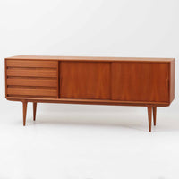 Gunni Omannがデザインし、Omann Jun社によって製造されたModel 18は、洗練されたラインと実用性を兼ね備えた北欧ビンテージの名作サイドボードです。チーク材の美しい木目が前面に広がり、引き出しとスライド扉の組み合わせが高い収納力を実現しています。特に繊細に削り出された取手や先細りの脚部が、軽やかで彫刻的な印象を与え、空間を上質に演出します。幅200cmのサイズ感は、リビングやダイニングの主役として活躍する存在感ある一台です。