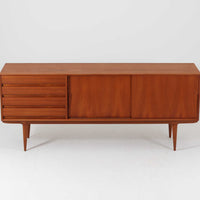 Gunni Omannがデザインし、Omann Jun社によって製造されたModel 18は、洗練されたラインと実用性を兼ね備えた北欧ビンテージの名作サイドボードです。チーク材の美しい木目が前面に広がり、引き出しとスライド扉の組み合わせが高い収納力を実現しています。特に繊細に削り出された取手や先細りの脚部が、軽やかで彫刻的な印象を与え、空間を上質に演出します。幅200cmのサイズ感は、リビングやダイニングの主役として活躍する存在感ある一台です。
