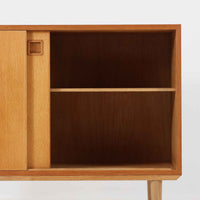 オーク材の美しい木目と柔らかな色味が魅力のこちらのボードは、幅120cmと使い勝手の良いサイズ感で、北欧ビンテージらしい洗練された佇まいが魅力です。内部には可動棚とフェルト付きの浅型引き出しが備えられ、日用品や小物の整理に最適。彫り込み式の取手やテーパードレッグなど、細部にまで配慮された上質な造りが光ります。販売店ラベルとして「Nr. Uttrup Boligmontering」が残されており、当時の流通背景を感じさせる一点物のアイテムです。