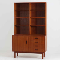 チーク材を使用したスウェーデン製のディスプレイシェルフです。上段は収納力のある可動棚付きのオープンスペース、下段には引き出し4杯と扉収納が備えられており、実用性とデザイン性を兼ね備えた構成です。シンプルな取手の意匠や直線的な脚部が北欧家具らしい洗練を感じさせ、経年により深みを増したチークの色合いが空間に温かみを与えます。リビングや書斎での本や雑貨の収納・展示におすすめのビンテージアイテムです。