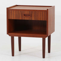 チーク材の豊かな木目が美しいこちらのサイドチェストは、北欧ビンテージらしい端正なデザインと実用性を兼ね備えた一台です。天板の縁には立ち上がりが施され、小物の転落を防ぐ工夫がされています。引き出し下のオープンスペースは本や雑貨の収納に便利で、ソファやベッドサイドに置けばサイドテーブルとしても活躍します。丸みを帯びた持ち手やテーパードレッグが柔らかな印象を与え、経年変化による木肌の深みが空間に温もりを添える逸品です。