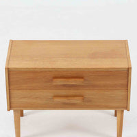 オーク材を用いたこちらの2段チェストは、北欧ビンテージ家具らしいシンプルかつ機能的なデザインが魅力です。直線的なフォルムに対し、細身でテーパーのかかった脚部が軽やかな印象を与え、空間を圧迫せずに置くことができます。幅60cmとコンパクトながら、2杯の引き出しは日用品や小物収納に便利で、ベッドサイドやリビングの補助収納としても最適です。経年により深みを増したオークの木肌が、温もりある雰囲気を演出する一点物です。