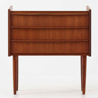 チーク材を使用したデンマーク製の3段チェストで、直線的な本体に対して丸みを帯びた引き手と細身のテーパードレッグが柔らかな印象を与え、さらに天板左右に伸びた耳のような張り出しがデザインのアクセントとなり、北欧らしい優雅な佇まいが感じられます。引き出しは3杯備え、衣類や小物収納に適したサイズ感で、ベッドサイドやリビングの一角に置きやすく、花器や照明のディスプレイ台としても楽しめます。シンプルな造形ながら木目の美しさが際立ち、空間に温かみと落ち着きを添えてくれるビンテージ家具です。