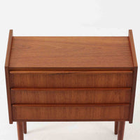 チーク材を使用したデンマーク製の3段チェストで、直線的な本体に対して丸みを帯びた引き手と細身のテーパードレッグが柔らかな印象を与え、さらに天板左右に伸びた耳のような張り出しがデザインのアクセントとなり、北欧らしい優雅な佇まいが感じられます。引き出しは3杯備え、衣類や小物収納に適したサイズ感で、ベッドサイドやリビングの一角に置きやすく、花器や照明のディスプレイ台としても楽しめます。シンプルな造形ながら木目の美しさが際立ち、空間に温かみと落ち着きを添えてくれるビンテージ家具です。