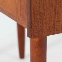 チーク材を使用したデンマーク製の3段チェストで、直線的な本体に対して丸みを帯びた引き手と細身のテーパードレッグが柔らかな印象を与え、さらに天板左右に伸びた耳のような張り出しがデザインのアクセントとなり、北欧らしい優雅な佇まいが感じられます。引き出しは3杯備え、衣類や小物収納に適したサイズ感で、ベッドサイドやリビングの一角に置きやすく、花器や照明のディスプレイ台としても楽しめます。シンプルな造形ながら木目の美しさが際立ち、空間に温かみと落ち着きを添えてくれるビンテージ家具です。