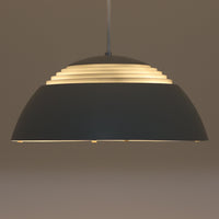 Arne JacobsenがLouis Poulsenのためにデザインした「AJ Royal 370」ペンダントライトです。直径37cmのサイズで、段階的に重なる上部シェードと、下面を覆うアクリルディフューザーによって光が柔らかく拡散し、均一で心地よい明かりを提供します。アルミとスチールの堅牢な構造に加え、シンプルかつモダンなフォルムが空間に洗練された印象を与えます。ダイニングやリビングに適した存在感ある照明で、北欧デザインを象徴するビンテージアイテムです。現行モデルでは展開されていないライトグレーのカラーは、当時のオリジナル品ならではの希少な仕様であり、コレクターズアイテムとしても高い価値を持っています。