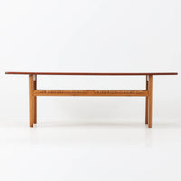 Hans J. WegnerがAndreas Tuckのために手掛けたAT10センターテーブルは、チーク材の天板にオーク材のフレーム、ラタン張りの棚板を組み合わせた、素材使いの美しさが際立つ北欧ビンテージの名作です。直線を基調とした端正なプロポーションの中に、天板縁のやわらかな丸みや、軽やかに抜け感を生むラタン棚が配され、視覚的にも圧迫感のない佇まいに仕上げられています。幅160cmのゆとりあるサイズはリビングの主役として使いやすく、雑誌や小物を収納できる実用性も備え、経年変化により深みを増した木肌が空間に上質な落ち着きをもたらします。なお、天板には反りがあり、大きめのへこみも見られるため、その点を考慮した価格設定としておりますが、ご使用には問題なく、ビンテージならではの風合いとしてご理解いただける方におすすめの一台です。