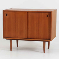 こちらの小ぶりなボードは、温かみのあるチーク材を用いたすっきりとした佇まいが魅力の北欧ビンテージ家具で、スライド扉を開くと左右に棚板付きの収納スペースが現れ、日用品や小物を効率よく整理できます。直線的な箱型の構造に、細身でテーパーの効いた脚部が軽やかな印象を添え、コンパクトながら存在感のある設計が空間に落ち着いたアクセントをもたらします。経年で深まった木肌の質感も味わい深く、リビングや玄関脇のサイド収納としても取り入れやすい一台です。