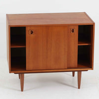 こちらの小ぶりなボードは、温かみのあるチーク材を用いたすっきりとした佇まいが魅力の北欧ビンテージ家具で、スライド扉を開くと左右に棚板付きの収納スペースが現れ、日用品や小物を効率よく整理できます。直線的な箱型の構造に、細身でテーパーの効いた脚部が軽やかな印象を添え、コンパクトながら存在感のある設計が空間に落ち着いたアクセントをもたらします。経年で深まった木肌の質感も味わい深く、リビングや玄関脇のサイド収納としても取り入れやすい一台です。