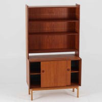 スウェーデン製のチーク材とオーク材を組み合わせたディスプレイシェルフです。幅90cmと程よいサイズ感で、上部の棚は書籍やオブジェの展示に適し、下部の引き戸収納は日用品や小物をすっきりと収めることができます。丸みを帯びた取っ手が温かみのあるアクセントとなり、北欧家具らしいシンプルで実用的なデザインを際立たせています。経年によって深みを増したチークの木目とオークのコントラストが上品な存在感を放ち、リビングや書斎の空間に落ち着いた雰囲気を添えるビンテージ家具です。