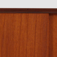 スウェーデン製のディスプレイシェルフです。上段には3段のオープンシェルフが設けられ、本や雑貨、オブジェなどを美しくディスプレイできます。下段にはスライド扉収納が備わり、内部の棚板によって整理しやすく、見せたくない収納にも便利です。チーク材の温もりある木目と、丸い掘り込み取っ手の柔らかなデザインが印象的で、北欧家具らしい穏やかな雰囲気を演出します。背面のグリーンの化粧板が空間に個性を添え、飾るアイテムをより際立たせます。本体にはチーク材、脚部にはオーク材が使用されており、素材のコントラストも魅力のひとつです。下段収納部の棚板にわずかな反りがありますが、裏返して使用することで目立たなくなります。軽やかな佇まいと実用性を兼ね備えた、北欧ビンテージらしい一台です。