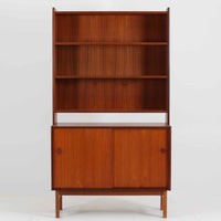 スウェーデン製のディスプレイシェルフです。上段には3段のオープンシェルフが設けられ、本や雑貨、オブジェなどを美しくディスプレイできます。下段にはスライド扉収納が備わり、内部の棚板によって整理しやすく、見せたくない収納にも便利です。チーク材の温もりある木目と、丸い掘り込み取っ手の柔らかなデザインが印象的で、北欧家具らしい穏やかな雰囲気を演出します。背面のグリーンの化粧板が空間に個性を添え、飾るアイテムをより際立たせます。本体にはチーク材、脚部にはオーク材が使用されており、素材のコントラストも魅力のひとつです。下段収納部の棚板にわずかな反りがありますが、裏返して使用することで目立たなくなります。軽やかな佇まいと実用性を兼ね備えた、北欧ビンテージらしい一台です。