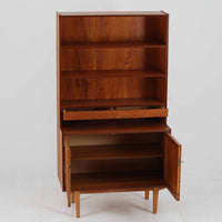 スウェーデン製のディスプレイシェルフです。上部は高さ調整可能な棚板が設けられており、書籍やオブジェの収納・展示に最適です。中央には小引き出しが2杯備わり、カトラリーや小物の整理にも便利。下部には扉付きの収納スペースがあり、日用品をすっきりと収納できます。素材にはチーク(本体)・オーク(脚の幕板)・ビーチ(脚)の3種の木材が用いられ、異なる木目のコントラストが美しい仕上がりです。北欧家具らしい温もりと実用性を兼ね備えた一台で、リビングやダイニング、書斎など幅広いシーンに調和します。