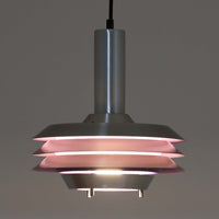 Carl Thoreがデザインし、Granhaga社より発表された直径28cmのペンダントライトです。アルミ製のシェードを幾重にも重ねた構造が特徴で、点灯すると隙間から柔らかな光が漏れ、独特の陰影と奥行きを生み出します。外側はシルバー、内側には紫がかった色味が施され、消灯時と点灯時で異なる表情を楽しめます。モダンでありながら温かみのある北欧らしい光のデザインは、ダイニングやリビングの照明として空間にアクセントを与えます。ビンテージならではの存在感を放つ希少な一品です。