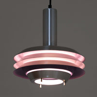 Carl Thoreがデザインし、Granhaga社より発表された直径28cmのペンダントライトです。アルミ製のシェードを幾重にも重ねた構造が特徴で、点灯すると隙間から柔らかな光が漏れ、独特の陰影と奥行きを生み出します。外側はシルバー、内側には紫がかった色味が施され、消灯時と点灯時で異なる表情を楽しめます。モダンでありながら温かみのある北欧らしい光のデザインは、ダイニングやリビングの照明として空間にアクセントを与えます。ビンテージならではの存在感を放つ希少な一品です。