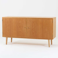 Poul Hundevadが手がけ、Hundevad & Coより製作されたキャビネットです。オーク材の端正な木目を活かしたフラットな扉構成と、細身のテーパードレッグが北欧モダンらしい軽やかさを演出します。内部は可動棚と可動式の浅型トレイを備え、書類や食器、小物まで用途に応じた整理が可能。天板下の構造や金物の収まりにも丁寧な作りが感じられ、長年の使用で深みを増した素材感がビンテージならではの魅力として空間に静かな存在感を添えます。