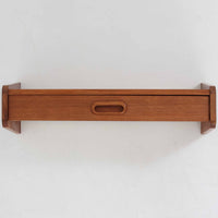 チーク材を用いたこちらのウォールシェルフは、直線的な棚板に両端の立ち上がりを設けた端正なフォルムが印象的な北欧ビンテージ家具です。前面には掘り込み式の取っ手を備えた引き出しが組み込まれており、見た目の軽やかさを保ちながら、実用的な収納力も確保しています。壁面を有効活用できる構造のため、デスク上やベッドサイド、玄関周りの小物収納としても取り入れやすく、側板は縁に向かって面取りが施され、厚みの変化によって立体感と柔らかな印象を与えています。経年により深みを増したチーク材の木目と丁寧な作りが、空間に静かな存在感を添える一台です。