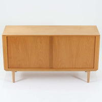 Poul Hundevadがデザインし、Hundevad & Coより製作された蛇腹仕様のサイドボードです。オーク材の穏やかで上品な木目を活かした外観に、横方向へ滑らかに開閉する蛇腹扉を組み合わせた、機能美あふれる一台。扉の開閉時に前後のスペースを取らない構造は、動線を妨げず実用性にも優れています。内部は棚板構成で収納力が高く、食器や書類、リビング周りの小物まで幅広く対応。細身の脚部が全体を軽やかに引き締め、経年によって深みを増した素材感が北欧ビンテージならではの静かな存在感を空間にもたらします。