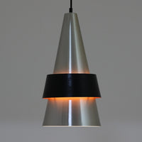 Jo HammerborgがFog & Morupのために手掛けた「Corona」は、直径22cmとコンパクトながら全高45cmと縦にしっかりとした存在感を持つアルミ×スチール製ペンダントライトで、上下に伸びる円錐形フォルムと中央のブラックバンドが彫刻的な印象を与える北欧ビンテージ品です。光源を囲む二重構造によって眩しさを抑えつつ下方向へ明かりを集中的に落とす設計となっており、ダイニングやカウンター上での1灯使用はもちろん、複数灯を並べても空間にリズムを生み出します。経年で深まった素材の風合いがハーマボーらしいモダンな佇まいを際立たせ、空間のアクセントとしても十分な存在感を発揮します。