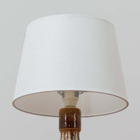 Soholm Keramik(スーホルム・セラミック)が手掛けたこちらのテーブルランプは、柔らかなクリーム色を基調にブラウンの縦流れ模様と円形のレリーフ装飾があしらわれた温かみある陶磁器ベースが魅力で、ふっくらと広がるフォルムが北欧ビンテージらしいやさしい存在感を放つ一台です。リネンシェード越しに灯る穏やかな光はベッドサイドやリビングの間接照明として心地よく空間を包み込み、経年で深まった釉薬の表情が一点物ならではの味わいを引き立てています。