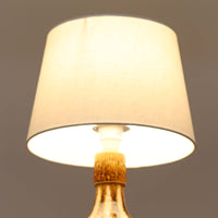 Soholm Keramik(スーホルム・セラミック)が手掛けたこちらのテーブルランプは、柔らかなクリーム色を基調にブラウンの縦流れ模様と円形のレリーフ装飾があしらわれた温かみある陶磁器ベースが魅力で、ふっくらと広がるフォルムが北欧ビンテージらしいやさしい存在感を放つ一台です。リネンシェード越しに灯る穏やかな光はベッドサイドやリビングの間接照明として心地よく空間を包み込み、経年で深まった釉薬の表情が一点物ならではの味わいを引き立てています。