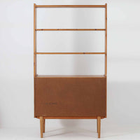スウェーデン製のこちらのオープンシェルフは、チーク材の温かな木目とオーク材の脚部を組み合わせた軽やかな佇まいが魅力で、上段の棚板と下段のスライド扉収納を備えた実用性の高い構成が特徴の北欧ビンテージ家具です。丸い掘り込み取手や細身のフレームなど繊細なディテールが空間を柔らかく彩り、本や雑貨、食器などを美しく見せながら整頓でき、経年で深まった木肌の風合いがインテリアに落ち着きと温もりを与える一台です。