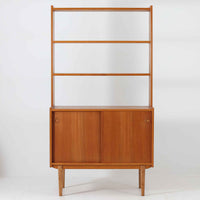 スウェーデン製のこちらのオープンシェルフは、チーク材の温かな木目とオーク材の脚部を組み合わせた軽やかな佇まいが魅力で、上段の棚板と下段のスライド扉収納を備えた実用性の高い構成が特徴の北欧ビンテージ家具です。丸い掘り込み取手や細身のフレームなど繊細なディテールが空間を柔らかく彩り、本や雑貨、食器などを美しく見せながら整頓でき、経年で深まった木肌の風合いがインテリアに落ち着きと温もりを与える一台です。