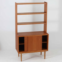 スウェーデン製のこちらのオープンシェルフは、チーク材の温かな木目とオーク材の脚部を組み合わせた軽やかな佇まいが魅力で、上段の棚板と下段のスライド扉収納を備えた実用性の高い構成が特徴の北欧ビンテージ家具です。丸い掘り込み取手や細身のフレームなど繊細なディテールが空間を柔らかく彩り、本や雑貨、食器などを美しく見せながら整頓でき、経年で深まった木肌の風合いがインテリアに落ち着きと温もりを与える一台です。