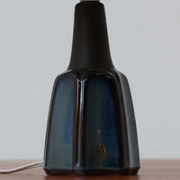 Einar JohansenがSoholm Keramik(スーホルム・セラミック)のためにデザインしたモデル1031は、深みのあるブルーの釉薬と立体的な面取りが織りなす陰影が美しい陶磁器ベースが特徴で、上部のマットな質感との対比がモダンで彫刻的な存在感を放つ北欧ビンテージ品です。プリーツシェードを透ける柔らかな灯りが落ち着いた雰囲気をつくり、ベッドサイドやリビングのアクセント照明として取り入れやすく、経年で深まった釉薬の表情が一点物ならではの魅力を一層引き立てています。