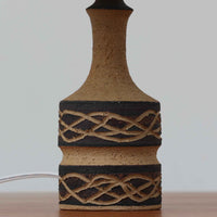 Jette HelleroeがAxellaのために手掛けたこちらのテーブルランプは、砂質の陶肌を生かした素朴で温かみのある質感と、ベース部分にぐるりと施された幾何学的なレリーフ装飾が印象的で、手仕事の風合いが際立つ北欧ビンテージ品です。すらりと立ち上がるシルエットが軽やかなバランスをつくり、リネンシェードを透ける柔らかな灯りがリビングやベッドサイドに落ち着いた雰囲気を添え、経年で深まった土味と装飾の陰影が一点物ならではの魅力を一層引き立てています。