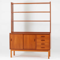 チーク材とビーチ材を組み合わせた北欧ビンテージらしい佇まいが魅力のディスプレイシェルフで、直線的で端正なフレーム構成と、木目の表情を活かした温かみのある質感が空間に落ち着きをもたらします。上部のオープン棚は器や書籍、オブジェの展示に適しており、下部には引き戸収納と引き出しを備えることで、見せる収納と隠す収納をバランス良く両立しています。脚付き構造により圧迫感が少なく、リビングやダイニング、書斎など幅広いシーンで活躍し、経年変化による色味の深まりも楽しめる一台です。