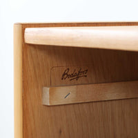 B. FridhagenがBodaforsのために手掛けたディスプレイシェルフは、端正なプロポーションと実用性を兼ね備えたスウェーデン製ビンテージ家具です。上部は抜け感のあるオープン棚構造で、器や書籍、オブジェなどを軽やかに飾ることができ、下部には4段の引き出しを備え、日常使いの収納にも対応します。オーク材ならではの明るく上品な木肌と、直線を基調とした構成が北欧らしい落ち着いた印象を生み、空間をすっきりとまとめてくれます。幅90cm、高さ122cmの小ぶりなサイズ感で、ダイニングやリビング、書斎など幅広いシーンに取り入れやすく、経年変化による木の表情も楽しめる一台です。