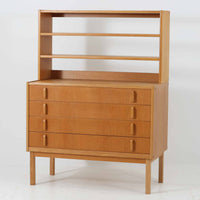 B. FridhagenがBodaforsのために手掛けたディスプレイシェルフは、端正なプロポーションと実用性を兼ね備えたスウェーデン製ビンテージ家具です。上部は抜け感のあるオープン棚構造で、器や書籍、オブジェなどを軽やかに飾ることができ、下部には4段の引き出しを備え、日常使いの収納にも対応します。オーク材ならではの明るく上品な木肌と、直線を基調とした構成が北欧らしい落ち着いた印象を生み、空間をすっきりとまとめてくれます。幅90cm、高さ122cmの小ぶりなサイズ感で、ダイニングやリビング、書斎など幅広いシーンに取り入れやすく、経年変化による木の表情も楽しめる一台です。