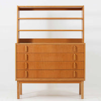 B. FridhagenがBodaforsのために手掛けたディスプレイシェルフは、端正なプロポーションと実用性を兼ね備えたスウェーデン製ビンテージ家具です。上部は抜け感のあるオープン棚構造で、器や書籍、オブジェなどを軽やかに飾ることができ、下部には4段の引き出しを備え、日常使いの収納にも対応します。オーク材ならではの明るく上品な木肌と、直線を基調とした構成が北欧らしい落ち着いた印象を生み、空間をすっきりとまとめてくれます。幅90cm、高さ122cmの小ぶりなサイズ感で、ダイニングやリビング、書斎など幅広いシーンに取り入れやすく、経年変化による木の表情も楽しめる一台です。