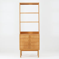 B. Fridhagenがデザインし、Bodafors社により1963年に製作されたスリムシェルフです。オーク材のやわらかな木目と明るい色合いが空間に温もりをもたらし、上部のオープン棚と下部の4段引き出しを備えた実用的な構成が魅力。幅63cmのすっきりとしたサイズで、書斎やリビングの一角にも取り入れやすい一台です。直線的で端正なフォルムと丁寧なつくりから、北欧ビンテージならではの機能美と経年の味わいを感じられる逸品です。