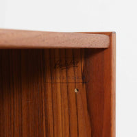 B.フリードホーゲンがデザインし、Bodafors社により製作された北欧ビンテージのディスプレイシェルフです。チーク材の美しい木目を活かした縦に伸びる端正なフレーム構成が印象的で、オープン棚と下部収納を組み合わせた実用性の高いデザインとなっています。上段は抜け感のある棚板構造で、書籍や器、オブジェを軽やかに飾ることができ、下段の引き戸収納は生活感の出やすい物をすっきりと収められます。脚付きによる軽快な佇まいと、経年変化によって深みを増した素材感が、空間に北欧ビンテージならではの落ち着いた存在感を与えます。