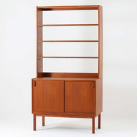 B.フリードホーゲンがデザインし、Bodafors社により製作された北欧ビンテージのディスプレイシェルフです。チーク材の美しい木目を活かした縦に伸びる端正なフレーム構成が印象的で、オープン棚と下部収納を組み合わせた実用性の高いデザインとなっています。上段は抜け感のある棚板構造で、書籍や器、オブジェを軽やかに飾ることができ、下段の引き戸収納は生活感の出やすい物をすっきりと収められます。脚付きによる軽快な佇まいと、経年変化によって深みを増した素材感が、空間に北欧ビンテージならではの落ち着いた存在感を与えます。