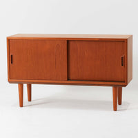 デンマークで製作されたチーク材のローボードです。幅100cmの小ぶりなサイズながら、温かみのある木目とシンプルな直線的フォルムが北欧らしい落ち着いた佇まいを演出します。前面には手掛け付きのスライド扉が備えられ、内部収納をすっきりと隠しながら使いやすさも確保。細身の脚部が軽やかな印象を与え、リビングや玄関、ベッドサイドなど様々な空間で活躍します。背板には壁掛け用の金具が取り付けられており、収納内部にはDIYによる木材の追加や小さな穴が見られますが、ビンテージ品ならではの味わいとして楽しめる一台です。