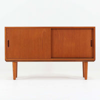 デンマークで製作されたチーク材のローボードです。幅100cmの小ぶりなサイズながら、温かみのある木目とシンプルな直線的フォルムが北欧らしい落ち着いた佇まいを演出します。前面には手掛け付きのスライド扉が備えられ、内部収納をすっきりと隠しながら使いやすさも確保。細身の脚部が軽やかな印象を与え、リビングや玄関、ベッドサイドなど様々な空間で活躍します。背板には壁掛け用の金具が取り付けられており、収納内部にはDIYによる木材の追加や小さな穴が見られますが、ビンテージ品ならではの味わいとして楽しめる一台です。