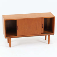 デンマークで製作されたチーク材のローボードです。幅100cmの小ぶりなサイズながら、温かみのある木目とシンプルな直線的フォルムが北欧らしい落ち着いた佇まいを演出します。前面には手掛け付きのスライド扉が備えられ、内部収納をすっきりと隠しながら使いやすさも確保。細身の脚部が軽やかな印象を与え、リビングや玄関、ベッドサイドなど様々な空間で活躍します。背板には壁掛け用の金具が取り付けられており、収納内部にはDIYによる木材の追加や小さな穴が見られますが、ビンテージ品ならではの味わいとして楽しめる一台です。